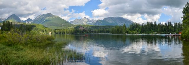 Serene panoramic view of an alpine lake surrounded by lush forests and towering mountains under a partly cloudy sky. Ideal for use in travel blogs, nature websites, and landscape photography portfolios, illustrating natural beauty and tranquility.