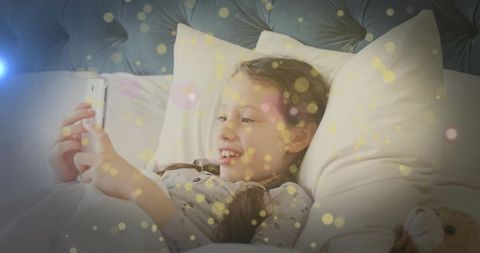 Smiling girl scrolling smartphone in cozy bed with tufted headboard, pajamas, teddy, bokeh