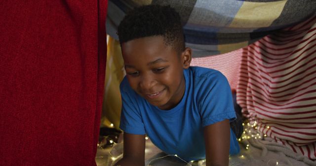 This image shows a young boy enjoying building and playing in a cozy blanket fort adorned with fairy lights. Perfect for promoting children's activities, creativity, playtime ideas, and indoor fun.