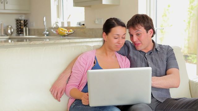 Couple seated on a comfortable sofa using a laptop for a video call in a relaxed living room environment. Ideal for illustrating concepts of modern technology, remote communication, online connectivity, social interaction from home, and digital engagement. Useful for content related to tech usage, lifestyle, and communication tools.