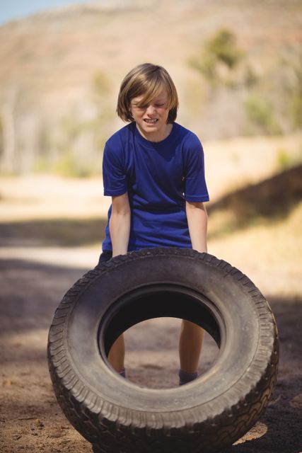 Young girl lifting a large tire during an outdoor boot camp obstacle course. She is showing determination and strength while exercising in a natural setting. Ideal for use in fitness, youth training, outdoor activities, and motivational content.