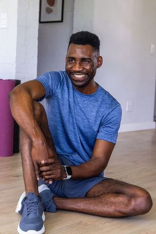 Smiling man sitting on hardwood floor holding knee after home workout, wearing smartwatch