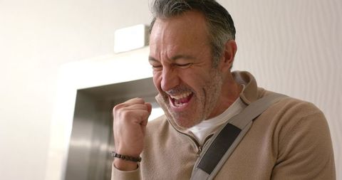 Mature man celebrating victory by elevator doors, raising clenched fist and smiling triumphantly
