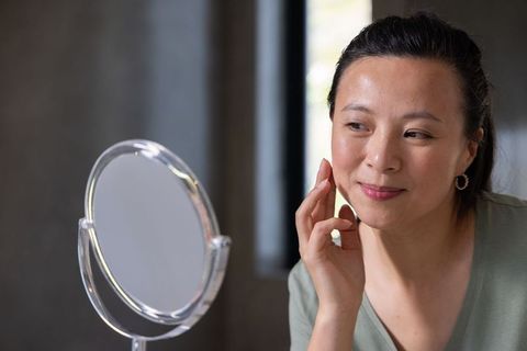 Mature woman touching cheek, studying face in vanity mirror by window, wearing sage top