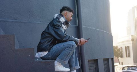 Young man sitting on urban steps wearing leather jacket and scarf using smartphone