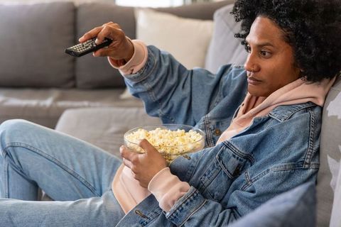 African american woman reclining on gray sofa holding popcorn and changing tv channel