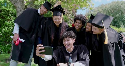 Diverse college graduates taking selfie on campus lawn celebrating graduation achievement