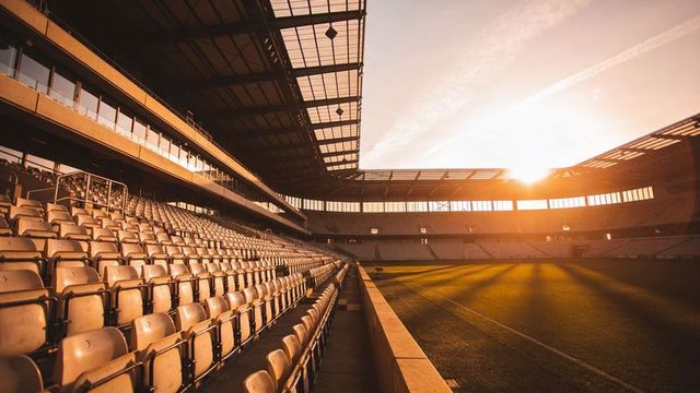 Empty stadium seats receding along sunlit football pitch, roof beams casting long shadows