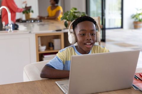 African american boy learning online with headphones at kitchen table, parents blurred