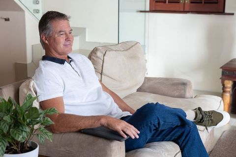Mature man relaxing on beige sofa holding tablet, casual jeans and white polo at home