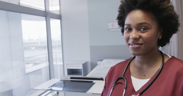 A nurse with an afro hairstyle is standing confidently in a hospital room, wearing a red uniform and a stethoscope around her neck. Her warm demeanor conveys professionalism and readiness to assist patients. Ideal for use in healthcare promotion, medical blogs, training materials, and articles focused on nursing and patient care.