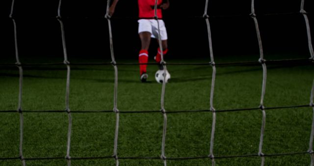 Depicts a soccer player in sports uniform preparing for goal kick. Net in foreground frames the action, emphasizing intensity and focus. Useful for promoting sporting events, athletic equipment, and fitness training programs, as well as illustrating articles focused on soccer strategies and sportsmanship.