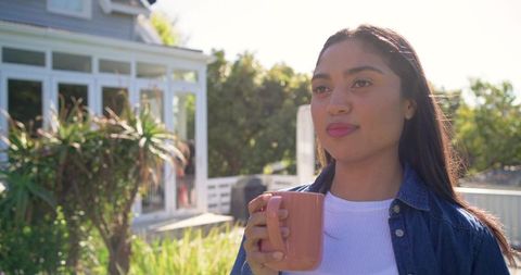 Young woman enjoying morning coffee on sunny backyard patio, holding ceramic mug, relaxing