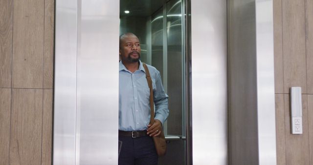 Businessman exiting a modern elevator in an office building carrying a shoulder bag. Ideal for business, corporate lifestyle, and commercial real estate promotions. Can be used to illustrate professional work environments or business attire.