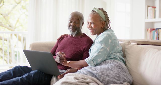 Happy senior african american couple holding hands and using laptop on couch in living room. Retirement, togetherness, communication, domestic life, technology and senior lifestyle, unaltered.
