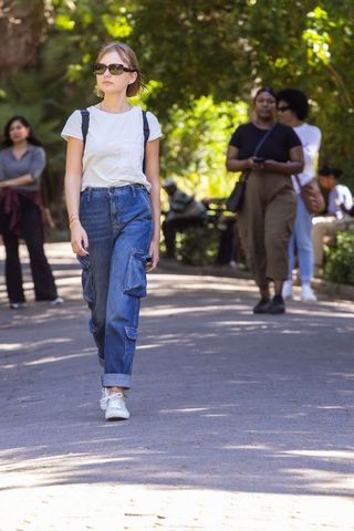 Young woman walking in sunny park wearing sunglasses, backpack, cargo jeans and sneakers