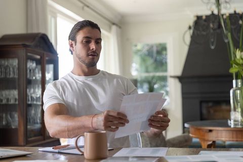 Man reviewing bills and documents at home workspace with coffee mug and notebook