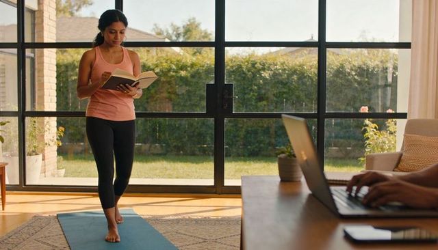 Woman reading book on yoga mat in sunlit living room with laptop nearby
