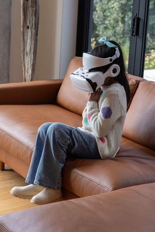 School-age girl sitting on leather sofa wearing vr headset using virtual reality in living room