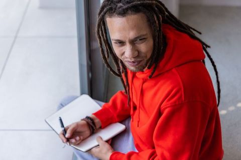 African american man writing in notebook by window wearing red hoodie, creative notetaking