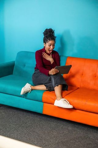 African american woman gasping while using tablet on vibrant teal and orange sofa