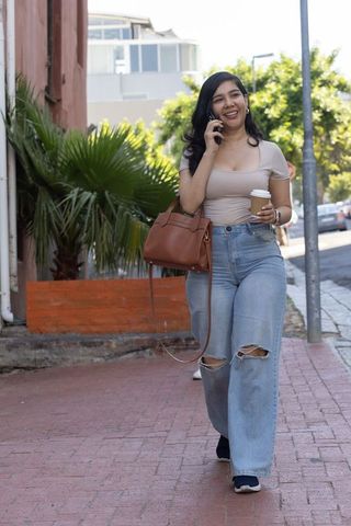Young woman walking city sidewalk talking on smartphone holding coffee and leather handbag