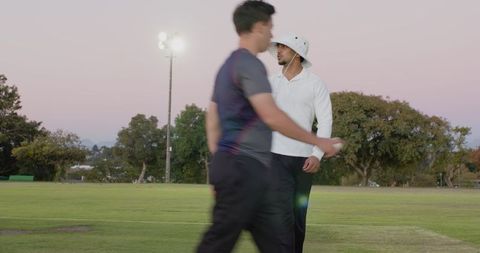 Indian coach surveying cricket field at dusk while player walking by holding ball