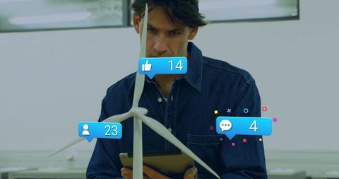 Engineer examining tabletop wind turbine prototype with tablet and social media alerts