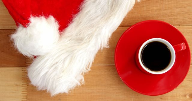 A red coffee cup sits next to a fluffy white Santa hat on a wooden surface, with copy space. The festive arrangement suggests a cozy holiday break or Christmas morning coffee.