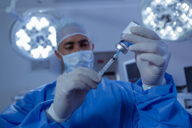 This image shows a surgeon in an operation room filling a syringe from an ampule. The surgeon is wearing protective gear, including gloves and a mask, ensuring a sterile environment. Can be used for medical-related content, hospital and healthcare promotions, or educational material concerning surgery procedures.