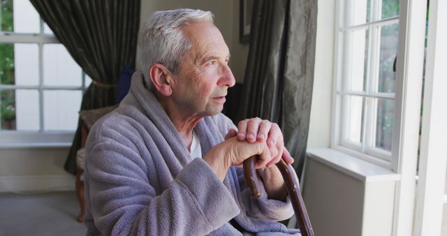 Elderly man sitting thoughtfully by a window, holding a cane and wearing a robe. Suitable for themes like retirement, senior care, solitude, reflection, and elderly living.