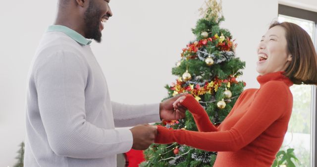 A joyful interracial couple laughing and enjoying time together in front of a beautifully decorated Christmas tree. Perfect for holiday greeting cards, festive season campaigns, promoting holiday happiness, or advertisements focused on love and togetherness during the holidays.