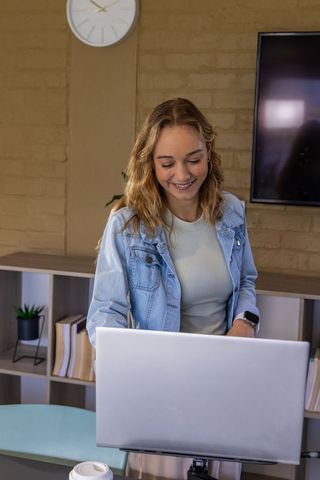 Young woman smiling and working on laptop at small office desk with coffee and monitor
