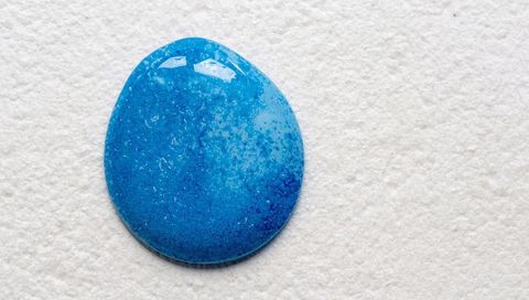 Glossy blue cabochon resting on coarse off-white textured surface, speckled highlights