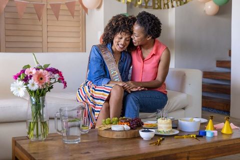 Black lesbian couple celebrating cozy home birthday, wearing sash and sharing cake