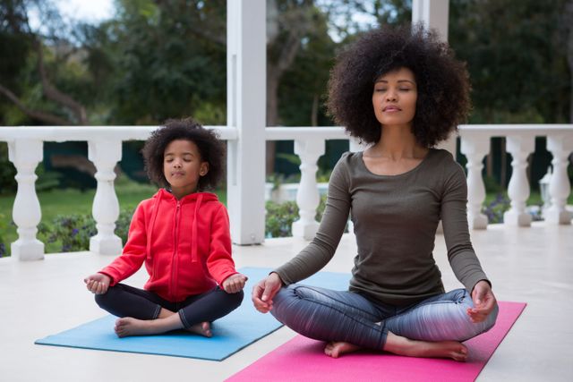 Mother and daughter meditating together on porch, practicing mindfulness and relaxation. Ideal for use in content related to family bonding, wellness, outdoor activities, and home life. Great for promoting calmness, mental health, and family relationships.
