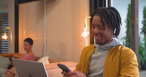 African american man using smartphone and laptop while partner relaxing on couch in cozy living room
