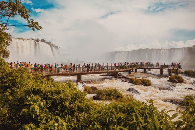 This depicts a large group of tourists on a bridge overlooking the majestic Iguazu Falls, which span the border between Brazil and Argentina. The powerful waterfalls and abundant greenery create a stunning landscape. This would be useful for materials related to travel, nature expeditions, eco-tourism, and South American destinations.