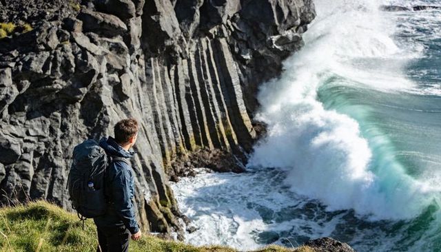 Hiker standing on cliff overlooking basalt columns and crashing turquoise ocean waves