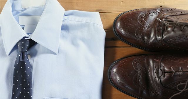 Formal attire with a blue dress shirt neatly folded alongside a pair of polished brown dress shoes and a blue polka dot tie on a wooden surface. Ideal for visuals related to men's fashion, office clothing, corporate style, dressing tips, and elegant wardrobes.