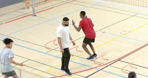 Diverse male athletes practicing agility hurdles in indoor gym with coach guiding drills