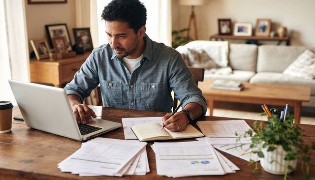 Reviewing finances and working from home at wooden desk with laptop, paperwork, plant
