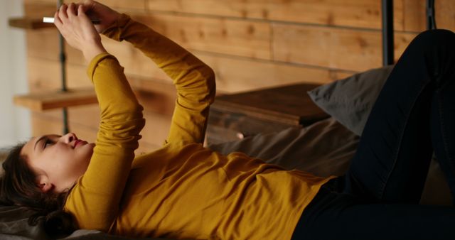Young woman lying on bed in yellow sweater, using smartphone with a wooden interior background. Perfect for illustrating modern lifestyle, relaxation, social media usage, or technology in everyday life.