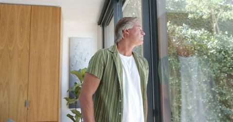 Mature man gazing through glass doors in modern home with sunlit reflection, contemplative moment