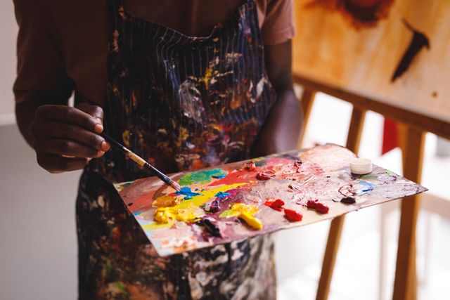 This image captures an African American male painter mixing vibrant colors on a palette in an art studio. Ideal for use in articles or advertisements related to creativity, art education, artistic inspiration, and the creative process. It can also be used for promoting art workshops, painting classes, and artistic events.