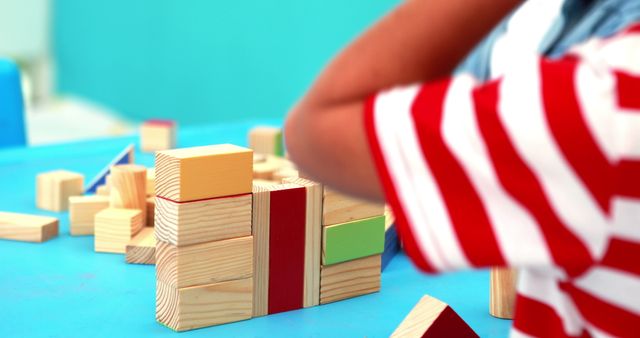 Child appears to be building a structure with colorful wooden blocks on blue table. Great for illustrating early childhood education, creativity, playful learning, and building cognitive skills. Can be used in advertisements for educational toys, parenting blogs, or kindergarten materials.