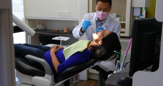 Dentist wearing a pink mask and black hair examining female patient's teeth sitting on dental chair. The clinic is equipped with modern dental tools and equipment. Suitable for use in materials related to dental care, oral health, healthcare services, or advertising a dental clinic.