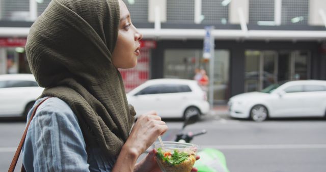 Happy biracial woman in hijab walking in city street eating takeaway salad. City living and healthy modern urban lifestyle.