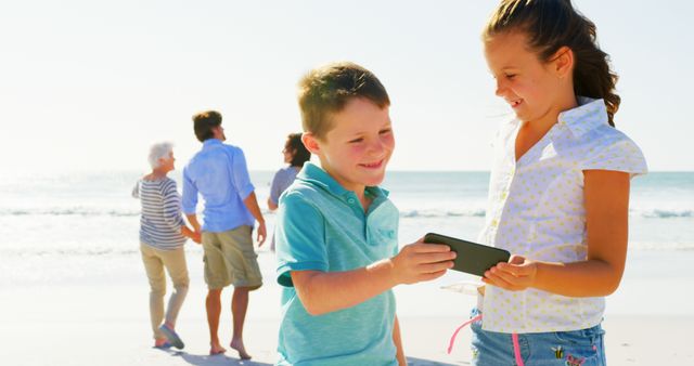 Kids standing on beach, smiling and enjoying looking at smartphone. Family with elders in background enjoying beach stroll. Great for advertisements, family travel blogs, summer vacation articles, and lifestyle projects.