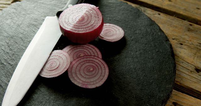 Freshly cut red onions lie on a rustic black slate, accompanied by a sharp kitchen knife. The thin slices highlight the texture and color of the onion, ideal for culinary content, food blogs, or advertisements related to cooking and kitchen supplies. This image can enhance recipes, ingredient showcases, or articles on food preparation.